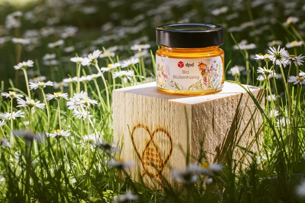Honigglas im Design von dpd auf einem Holzwürfel mit Zelenka-Brandzeichen in der Blumenwiese