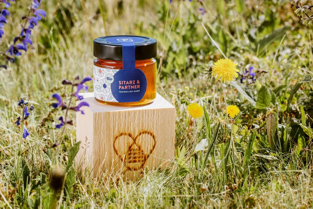 Honigglas im Design von Sitarz & Partner auf einem Holzwürfel mit Zelenka-Brandzeichen in der Blumenwiese